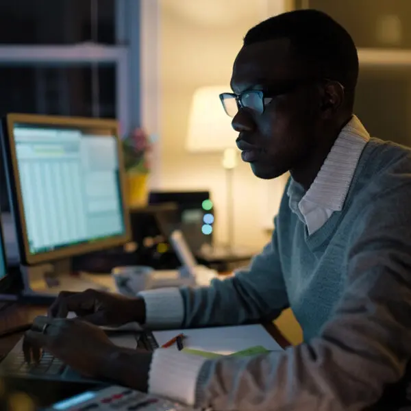 A focused man analyzing data on multiple computer screens in a dark room, highlighting the intensity and dedication required in a finance profession